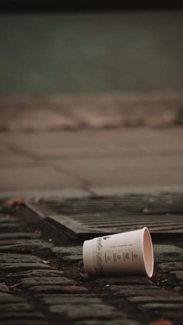 A middle-aged woman with short dark hair, dressed in a black T-shirt with a white graphic and dark trousers, is seen disposing of trash into a sleek, modern stainless steel public waste bin with a cyl
