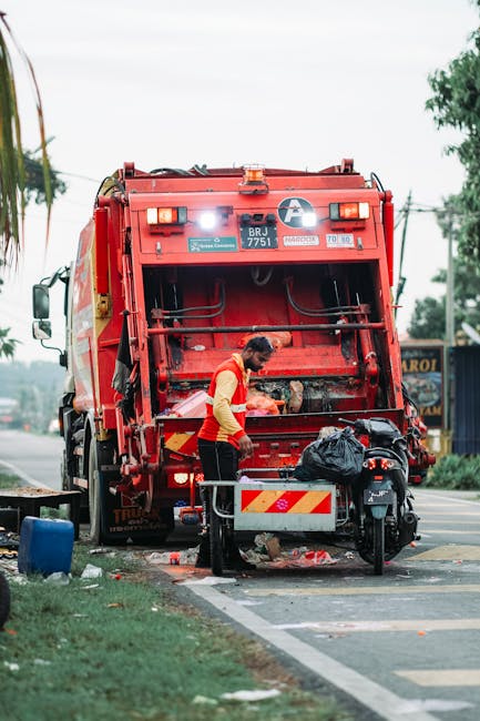 A pile of mixed household waste and rubbish, including cardboard boxes, plastic containers, black and grey storage bins, and various small household items, all scattered among dry, brown twigs and bra
