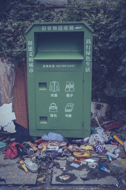A green old clothing recycling box situated outdoors against a background of dense foliage and trees, with a slightly weathered appearance. The box features Chinese characters and a phone number on it