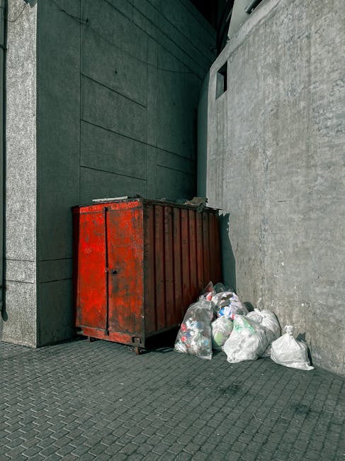 A large, weathered red metal skip with rust patches sits on a paved urban alleyway beside a concrete wall and a dark green building facade. The skip's surface appears rough with peeling paint and visi